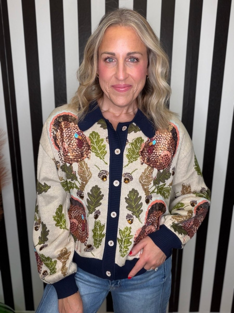 A woman wearing a cream-colored knit cardigan with a detailed autumn pattern of acorns, leaves, and large brown birds, paired with blue jeans, standing against a black and white striped background.