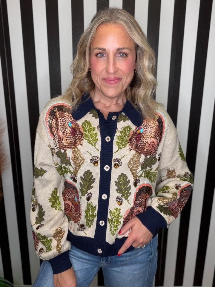 A woman wearing a cream-colored knit cardigan with a detailed autumn pattern of acorns, leaves, and large brown birds, paired with blue jeans, standing against a black and white striped background.