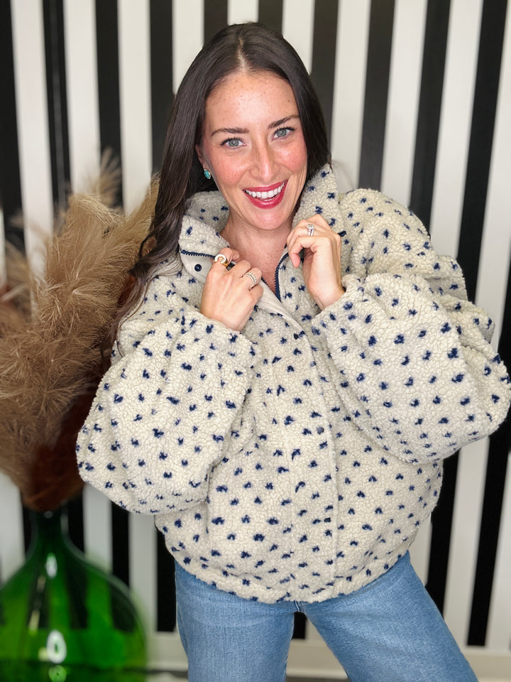 A woman wearing a cream sherpa fleece jacket with a navy blue speckled pattern, paired with light blue jeans, posing in front of a black and white striped background.