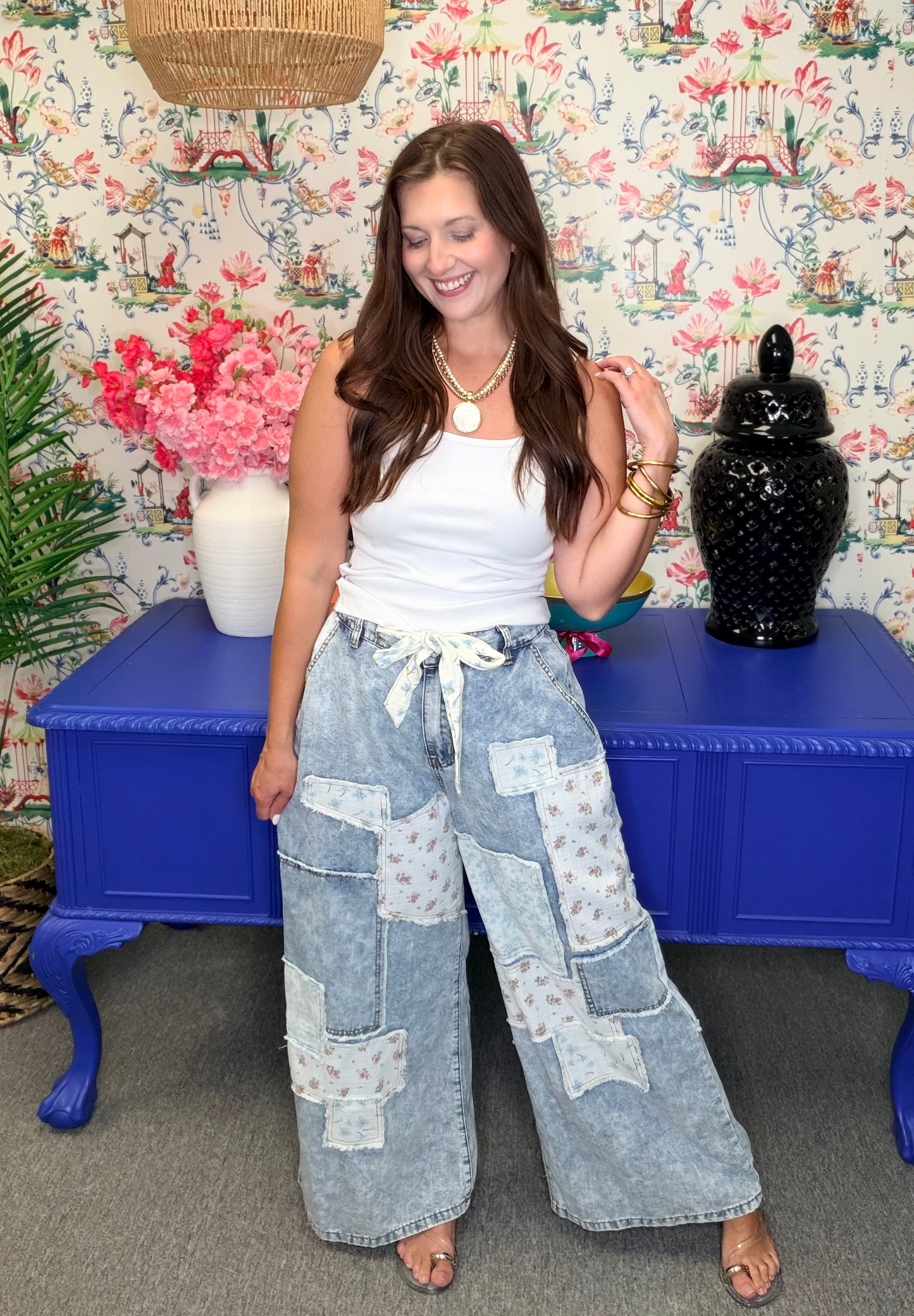 Woman wearing a white top and patchwork denim pants standing in front of a colorful floral-patterned wall.