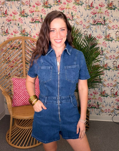 Woman wearing a blue denim romper standing in a room with decorative wallpaper and furniture.