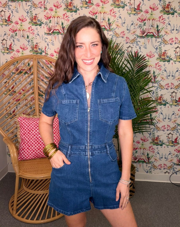 Woman wearing a blue denim romper standing in a room with decorative wallpaper and furniture.