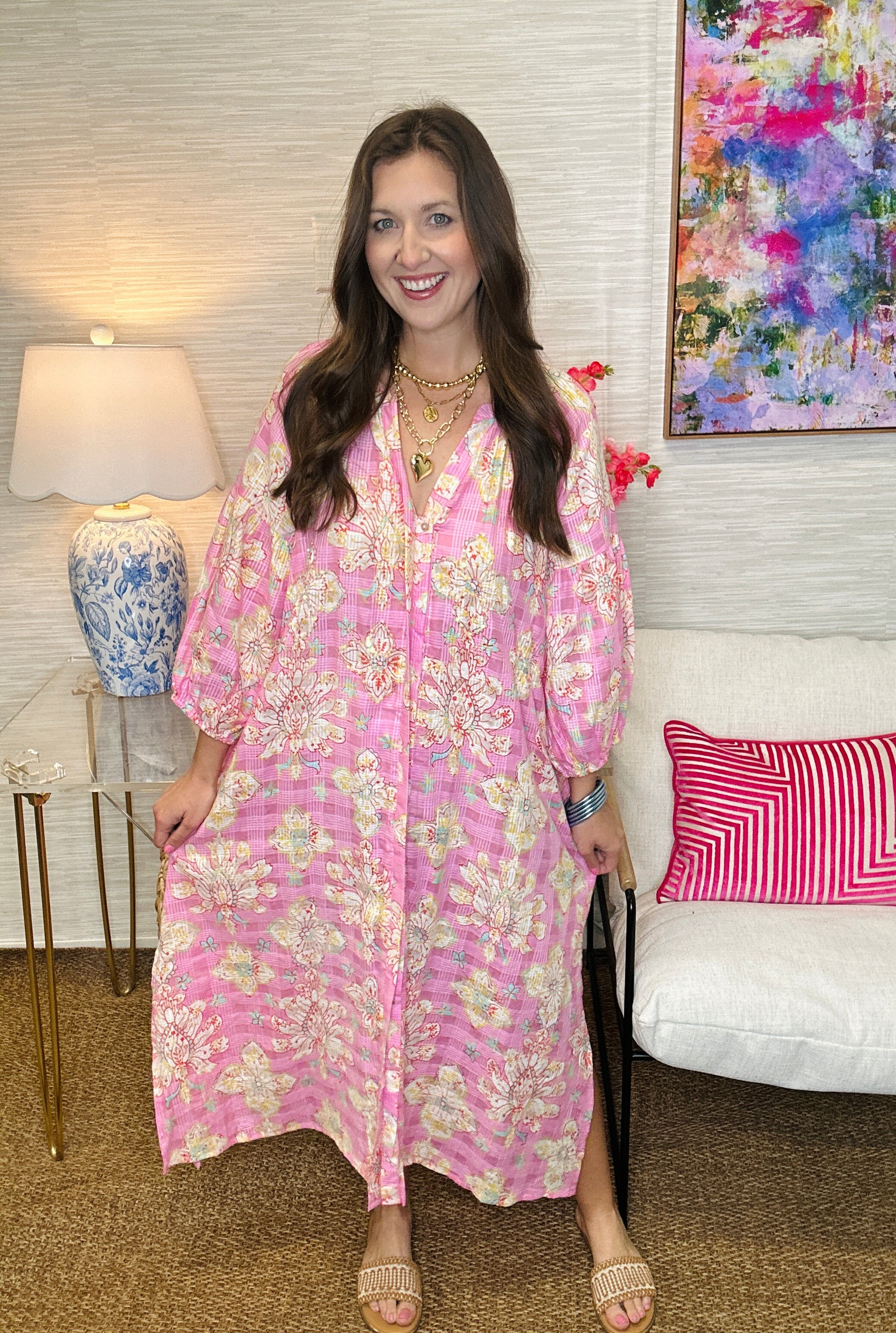 Woman in a pink floral dress standing in a room with a lamp and colorful painting.