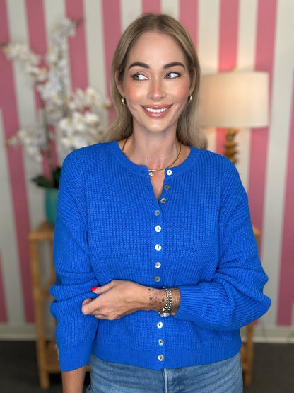 A woman wearing a bright royal blue waffle-knit button-up cardigan sweater, standing in front of a pink and white striped wall.