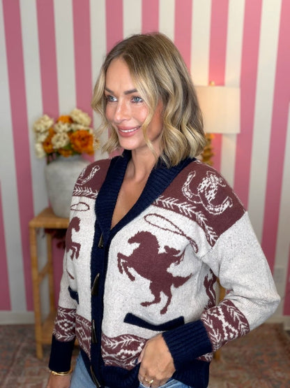 A woman wearing a patterned knit cardigan sweater with a horse motif, navy blue ribbed trim, and gold buttons, standing in front of a pink and white striped wall.