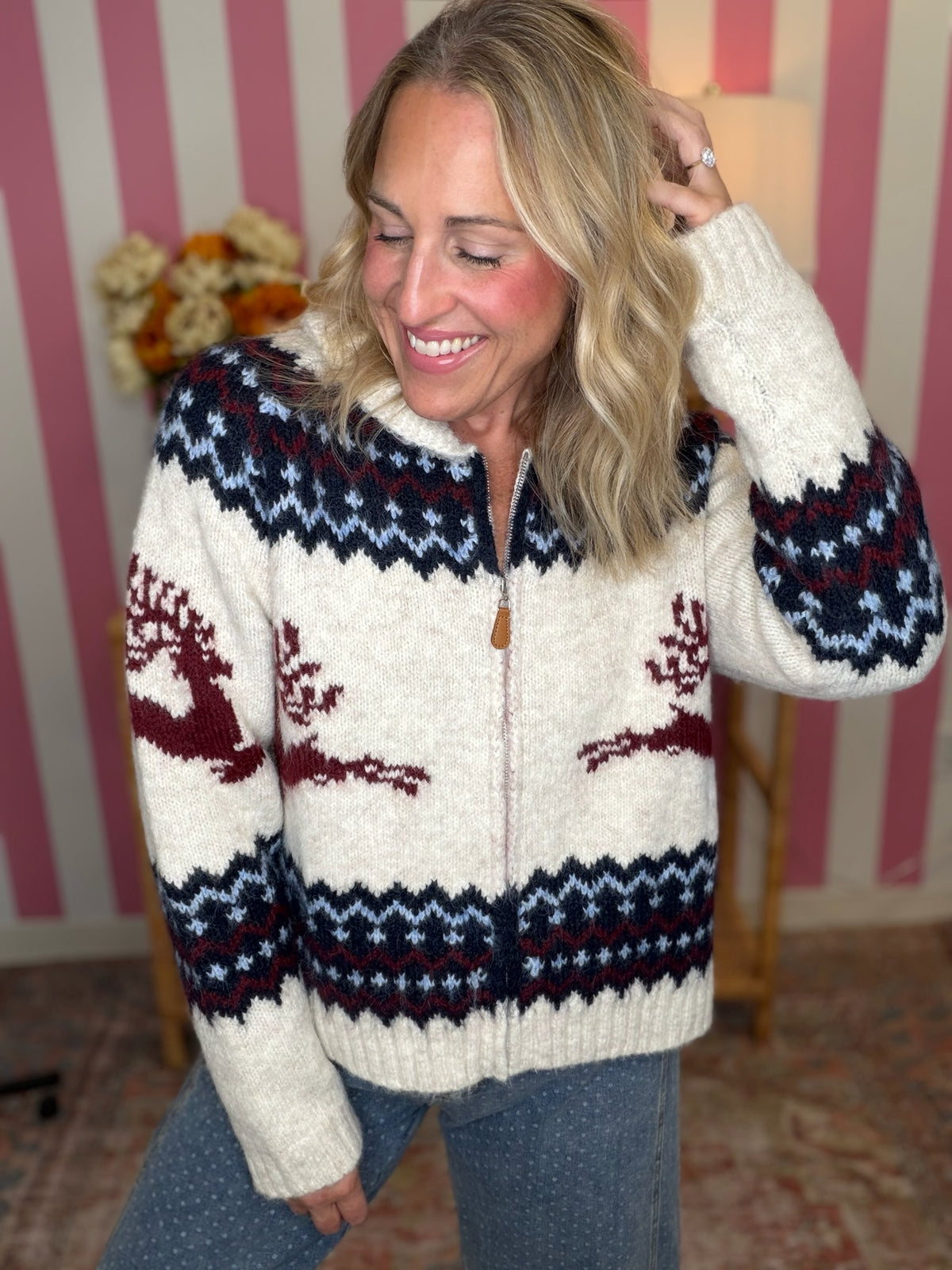 A woman wearing a cream-colored knit zip-up sweater with navy and burgundy Fair Isle patterns, including reindeer motifs, standing in front of a pink and white striped wall.