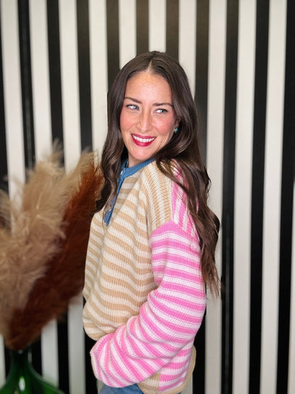 A woman with long brown hair smiles while wearing a color-block knit sweater with beige and white horizontal stripes on one side and pink and white stripes on the other, paired with a blue top underneath. She is standing in front of a black and white vertically striped background.