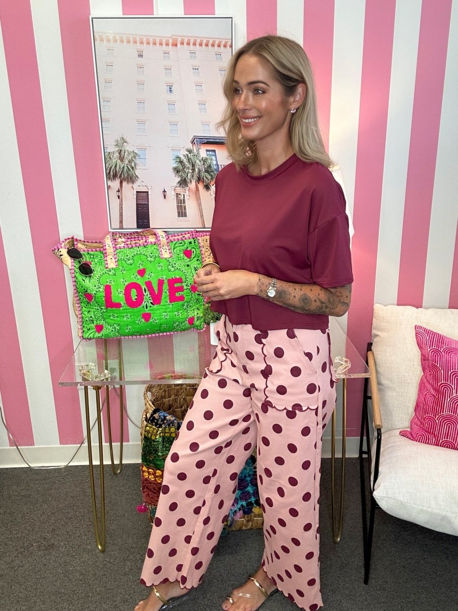 A woman stands in a room with pink and white striped walls, wearing a dark burgundy t-shirt and light pink wide-leg pants with dark burgundy polka dots. She is standing next to a clear glass table holding a bright green handbag with pink 'LOVE' lettering.