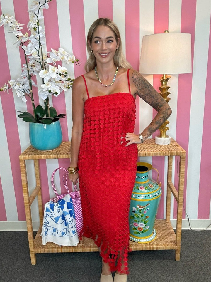 A woman wearing a bright red crochet knit dress stands in front of a pink and white striped wall, posing next to a wooden side table with a lamp, a blue vase with white flowers, and a colorful ceramic vase.