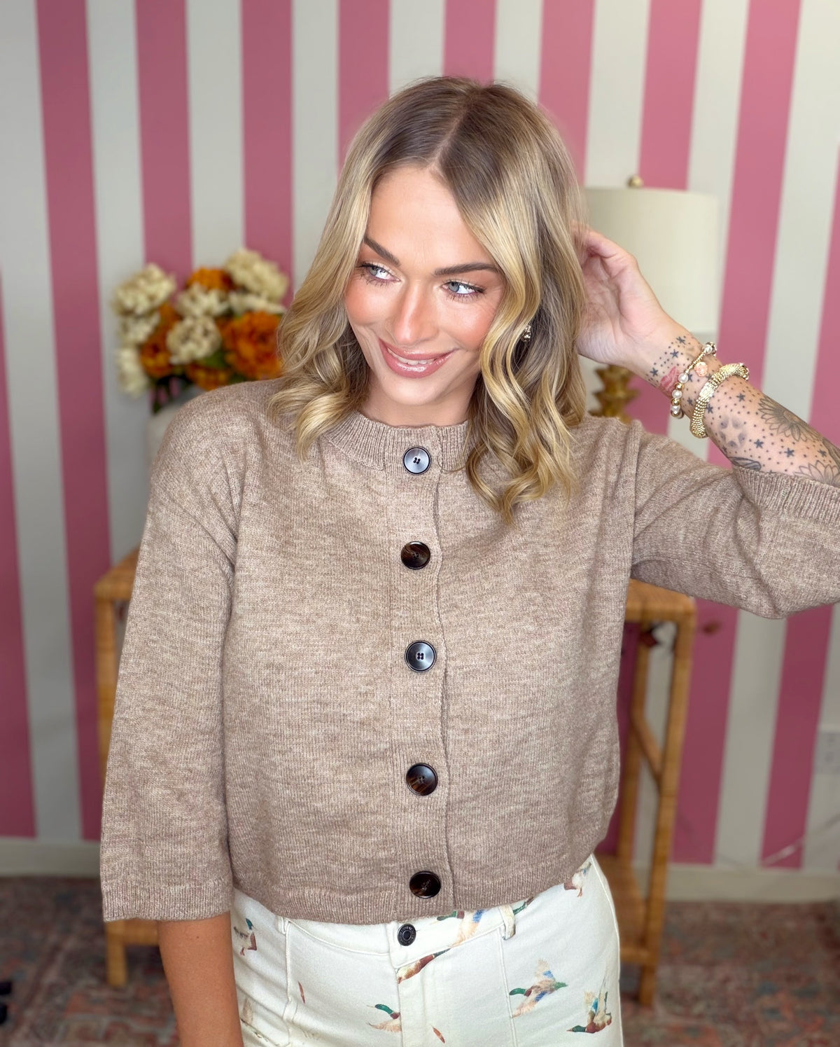 A smiling blonde woman wearing a beige knit cardigan with dark buttons, posed in front of a pink and white striped wall.