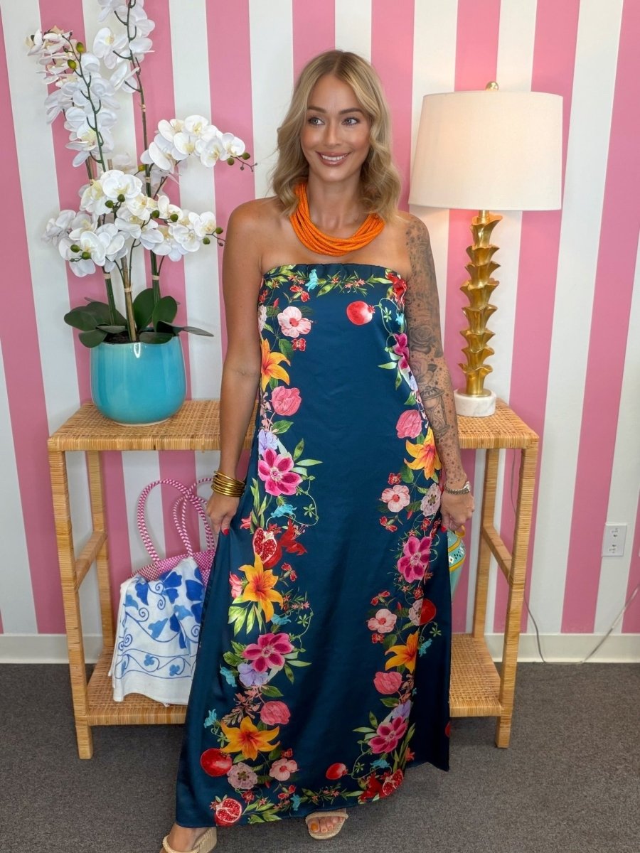 A woman modeling a strapless navy maxi dress with a vibrant multicolor floral print, styled with an orange necklace, standing in front of a pink and white striped wall.