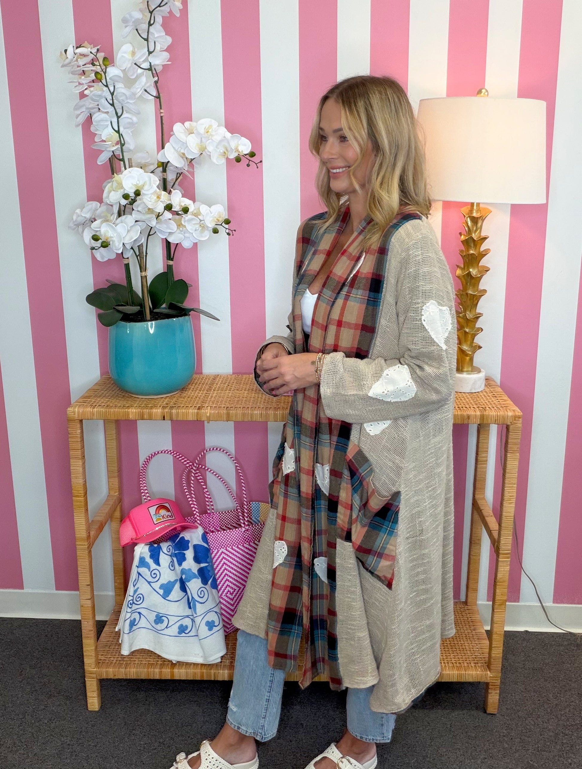A woman stands in a room with pink and white striped wallpaper, wearing a long beige cardigan with white heart patches over a plaid scarf and jeans. A wooden side table with a blue orchid pot and pink handbags is in front of her, along with a lamp.