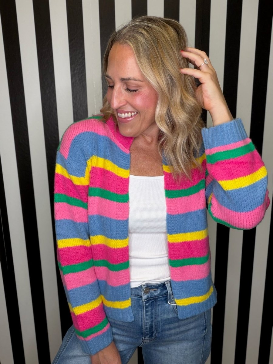 A woman wearing a colorful striped knit cardigan with pink, blue, yellow, and green horizontal stripes, paired with a white tank top and blue jeans, standing in front of a black and white striped wall.