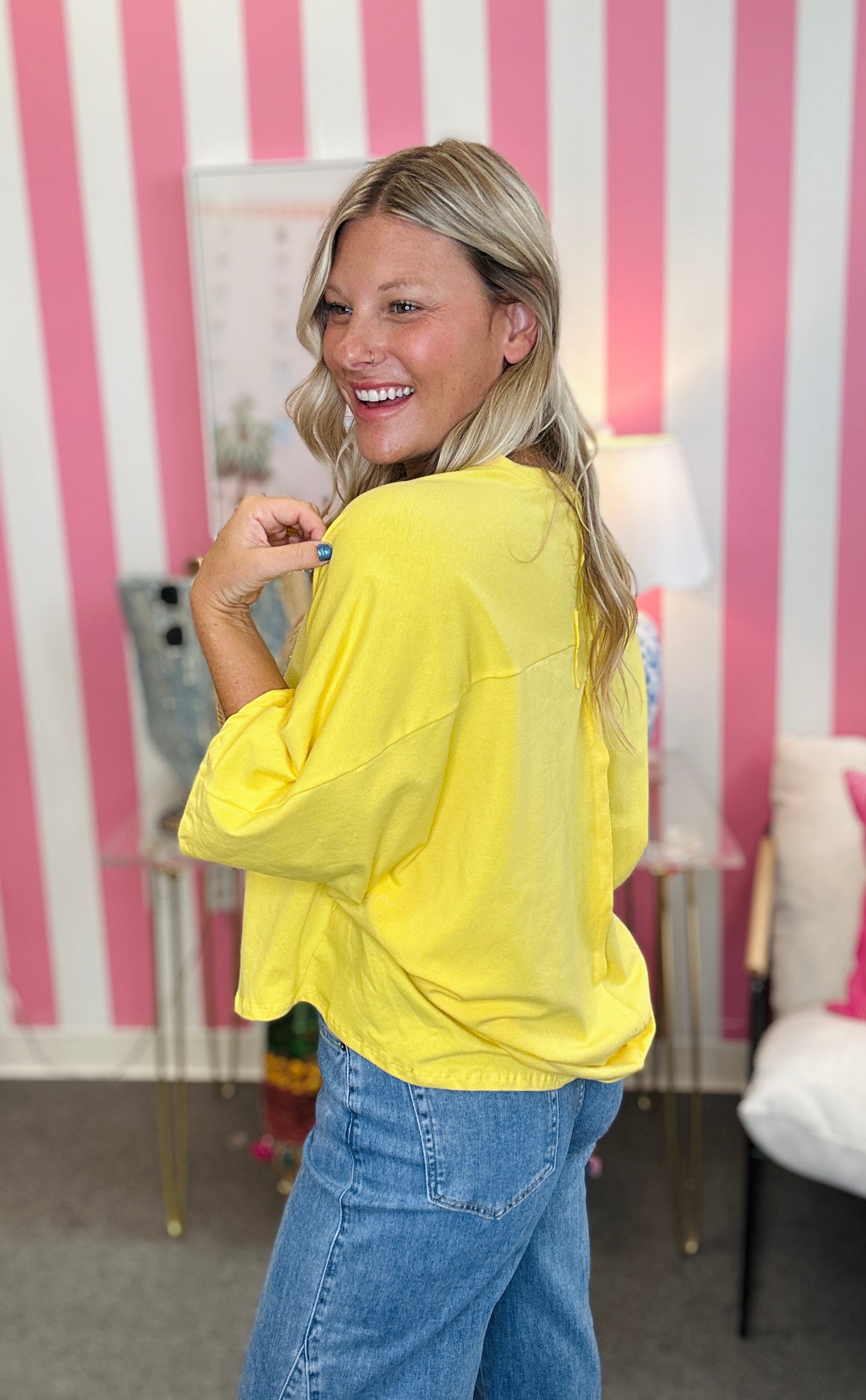 A woman with blonde hair smiling and looking back over her shoulder while wearing a bright yellow oversized long-sleeve top and blue jeans, standing in front of a pink and white striped wall.