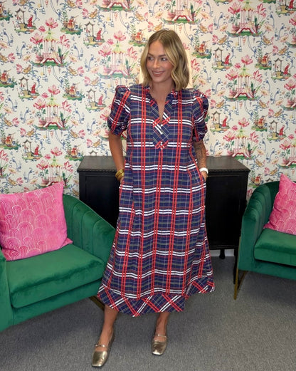 A woman stands in a room wearing a navy, red, and white plaid midi dress with puff sleeves. The background features a busy floral wallpaper, green velvet armchairs with pink pillows, and a black cabinet.