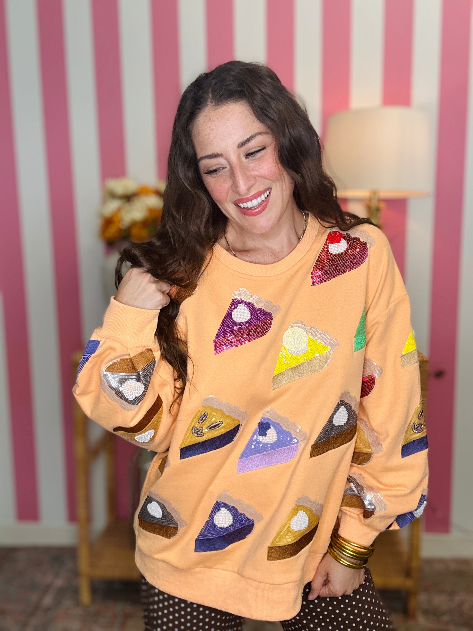 A woman smiling while wearing a peach-colored sweatshirt covered in sequined pie-slice patches, posed in front of a pink and white striped wall.