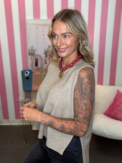 A woman with blonde hair and tattoos on her arms is wearing a beige knit sleeveless top and dark jeans, standing in front of a pink and white striped wall.