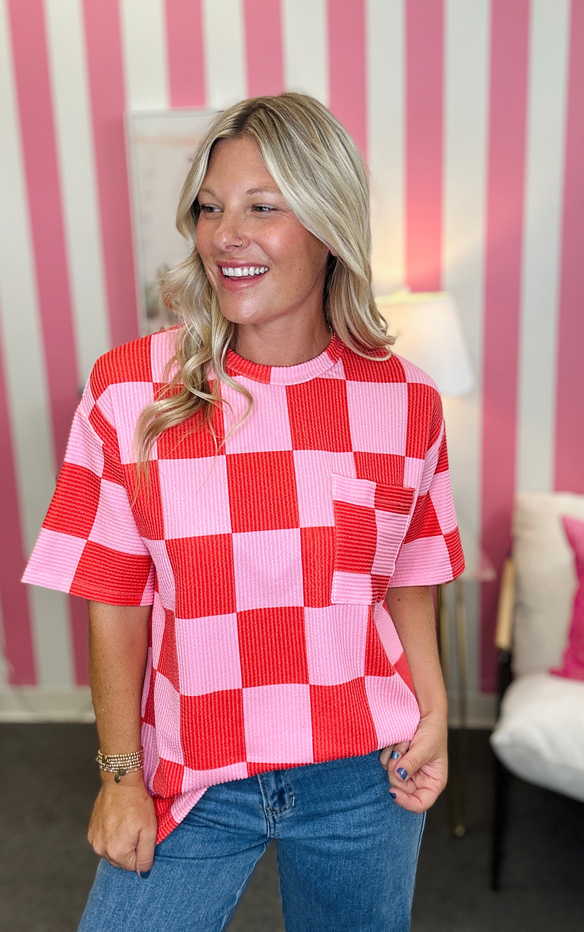 A woman wearing a pink and red checkered short-sleeve top with a chest pocket, paired with blue jeans, standing in front of a pink and white striped wall.