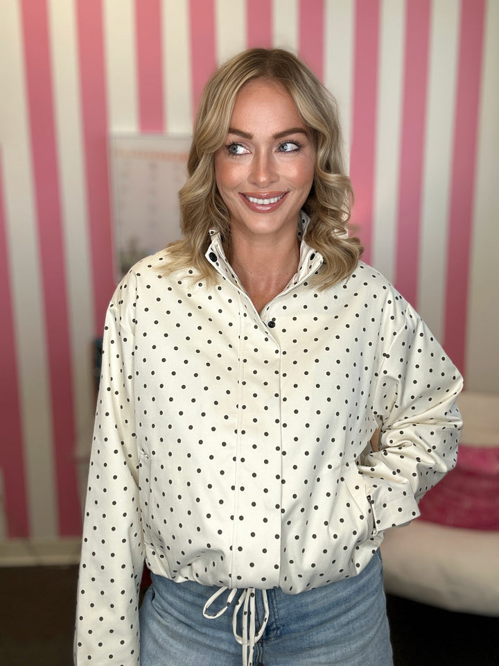 A woman with blonde hair wearing a cream-colored long-sleeve button-up shirt with black polka dots, paired with light blue jeans. She is standing in front of a pink and white striped wall.