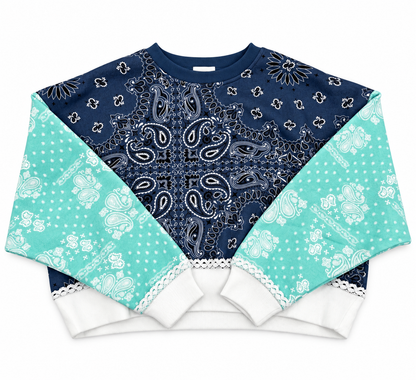 A cropped crewneck sweatshirt featuring a complex bandana print collage. The center panel is a dark navy blue paisley bandana print, while the sleeves are light aqua/mint green floral bandanas, finished with solid white ribbed cuffs and hem.