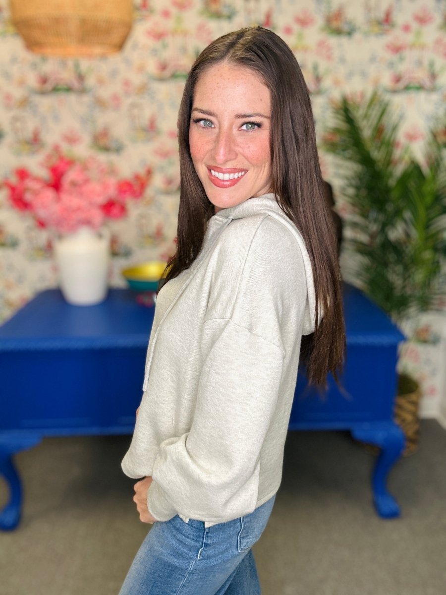 A woman with long brown hair smiling and looking back over her shoulder, wearing a light heather gray hoodie and blue jeans, standing in front of a floral wallpaper background with a blue table.