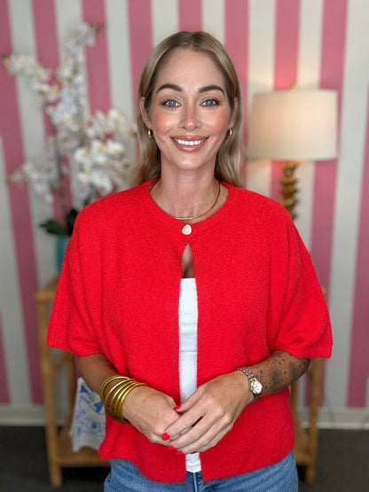A woman wearing a bright red short-sleeve knit top with a white inner layer and blue jeans, standing in front of pink and white striped wallpaper.