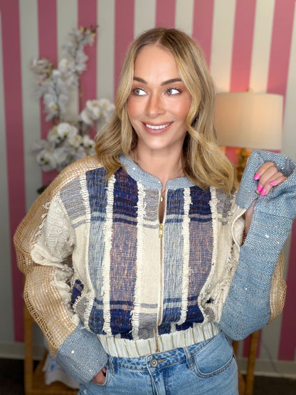 A woman wearing a cropped knit zip-up sweater with a plaid pattern in cream, navy, and gray, styled with light-wash blue jeans. She is standing in front of a pink and white striped wall with a lamp and flowers in the background.