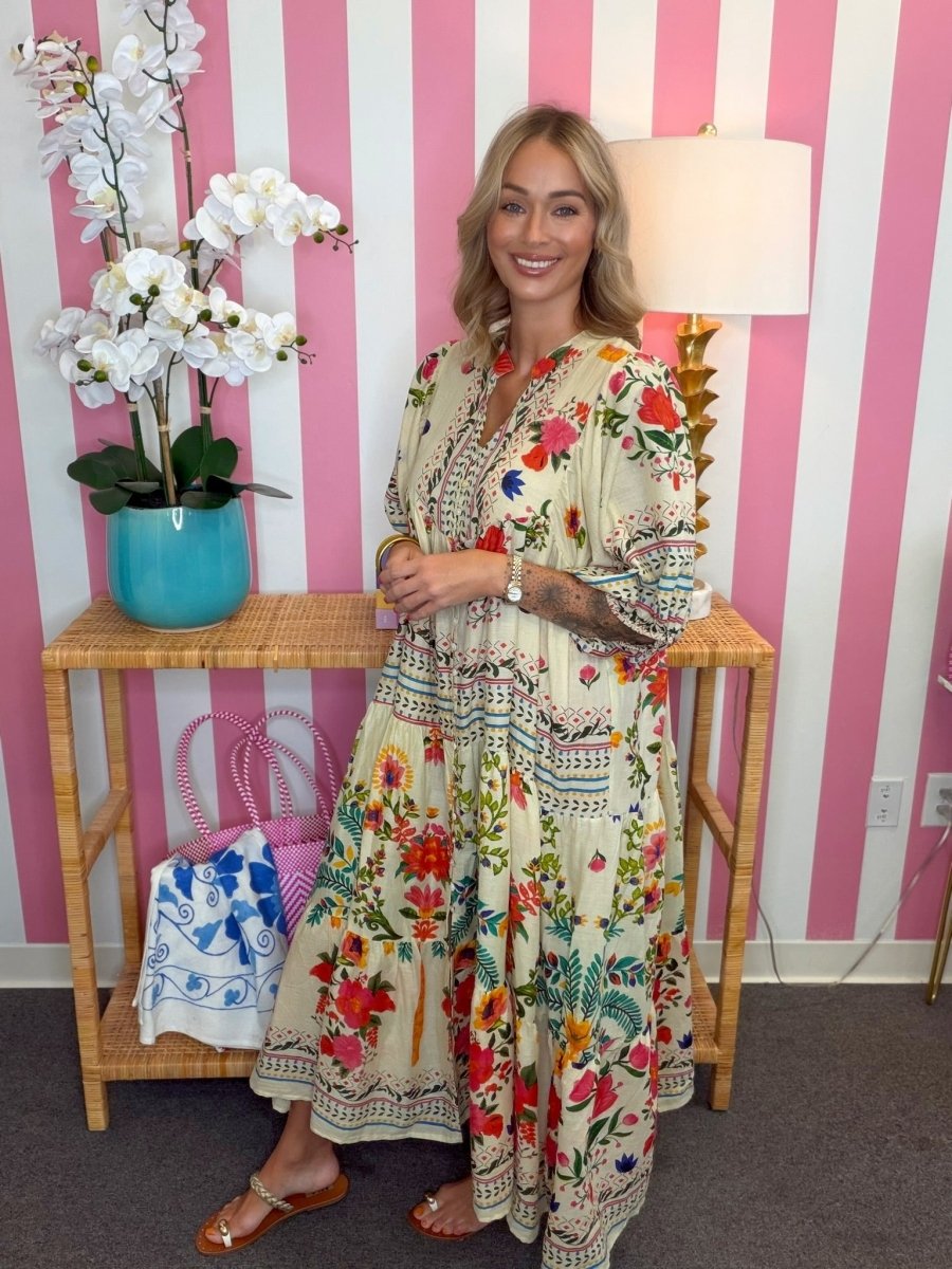 A woman wearing a bohemian-style maxi dress with a cream base and colorful floral print, standing in front of a pink and white striped wall next to a bamboo console table with a lamp and potted orchids.