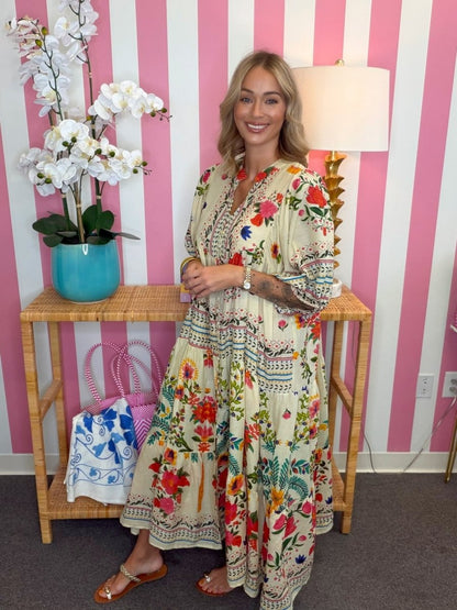 A woman wearing a bohemian-style maxi dress with a cream base and colorful floral print, standing in front of a pink and white striped wall next to a bamboo console table with a lamp and potted orchids.