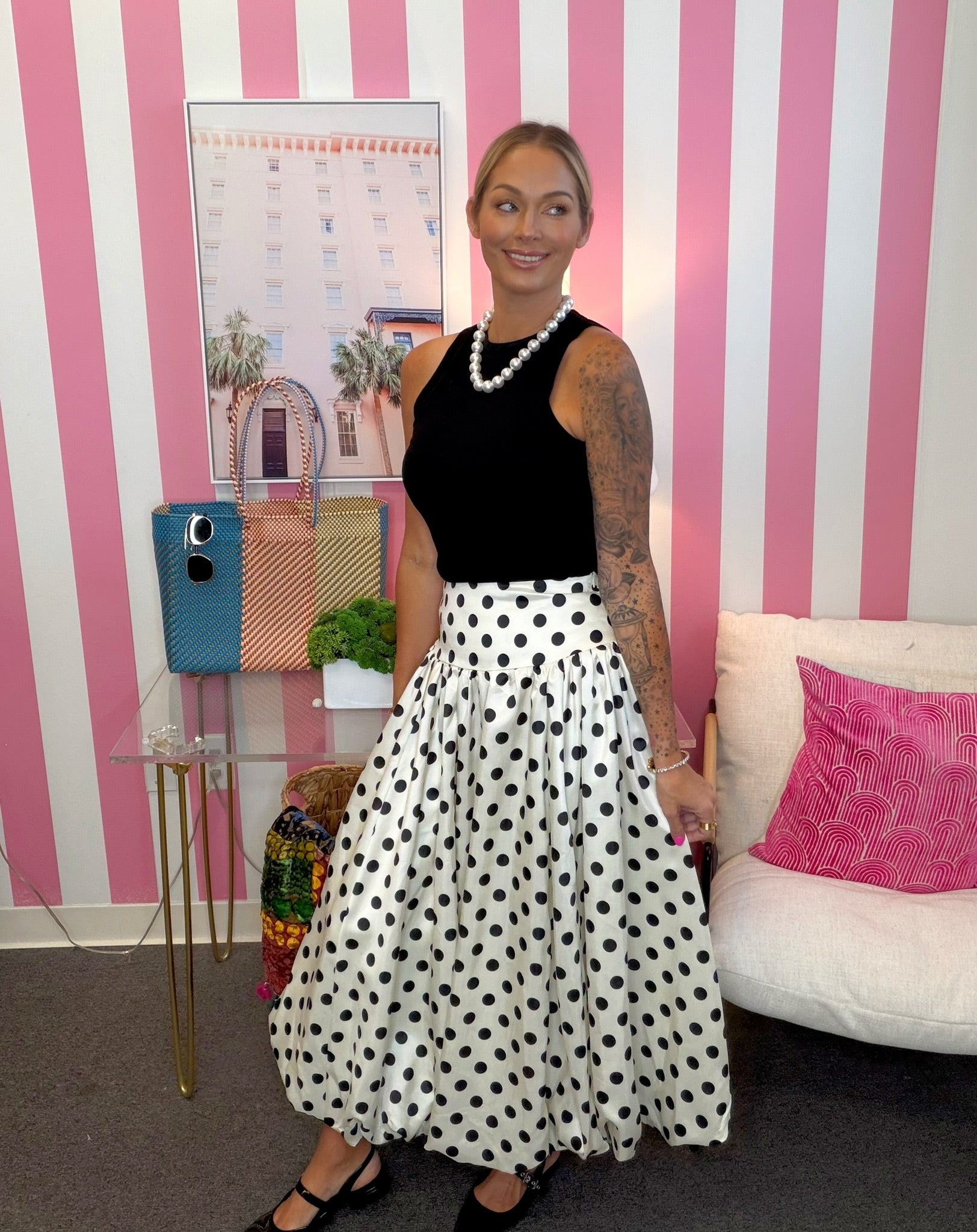 A woman stands in a room with pink and white striped walls, wearing a sleeveless black top and a white skirt with black polka dots. She is accessorized with a pearl necklace and black flat shoes.