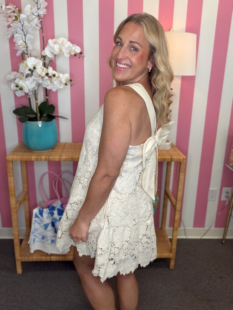 A woman wearing a sleeveless cream lace dress with a bow detail on the back, standing in front of a pink and white striped wall next to a bamboo side table with a blue orchid pot.