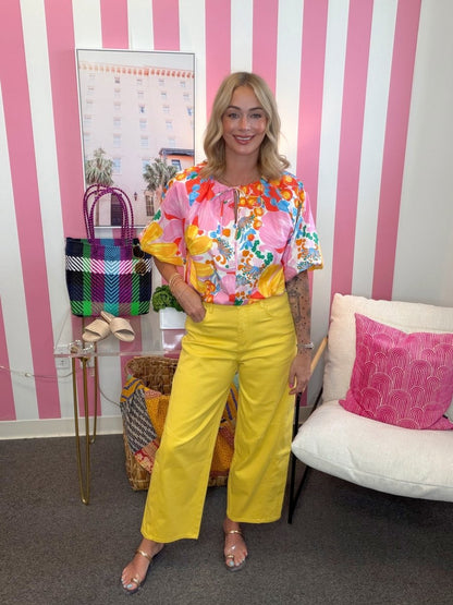 Woman in colorful floral blouse and yellow pants standing in a room with striped walls and furniture.