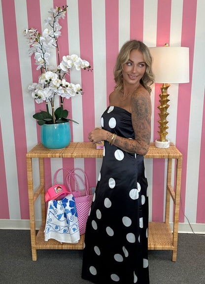 A woman wearing a black strapless maxi dress with large white polka dots stands in front of a pink and white striped wall, next to a wooden console table with a turquoise orchid pot and a lamp.