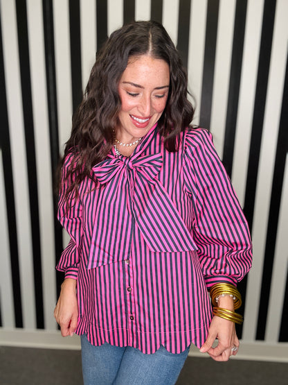 A woman wearing a pink and purple vertically striped blouse with a large bow at the collar, puff sleeves, and scalloped hem, paired with blue jeans and gold bracelets, standing in front of a black and white striped wall.