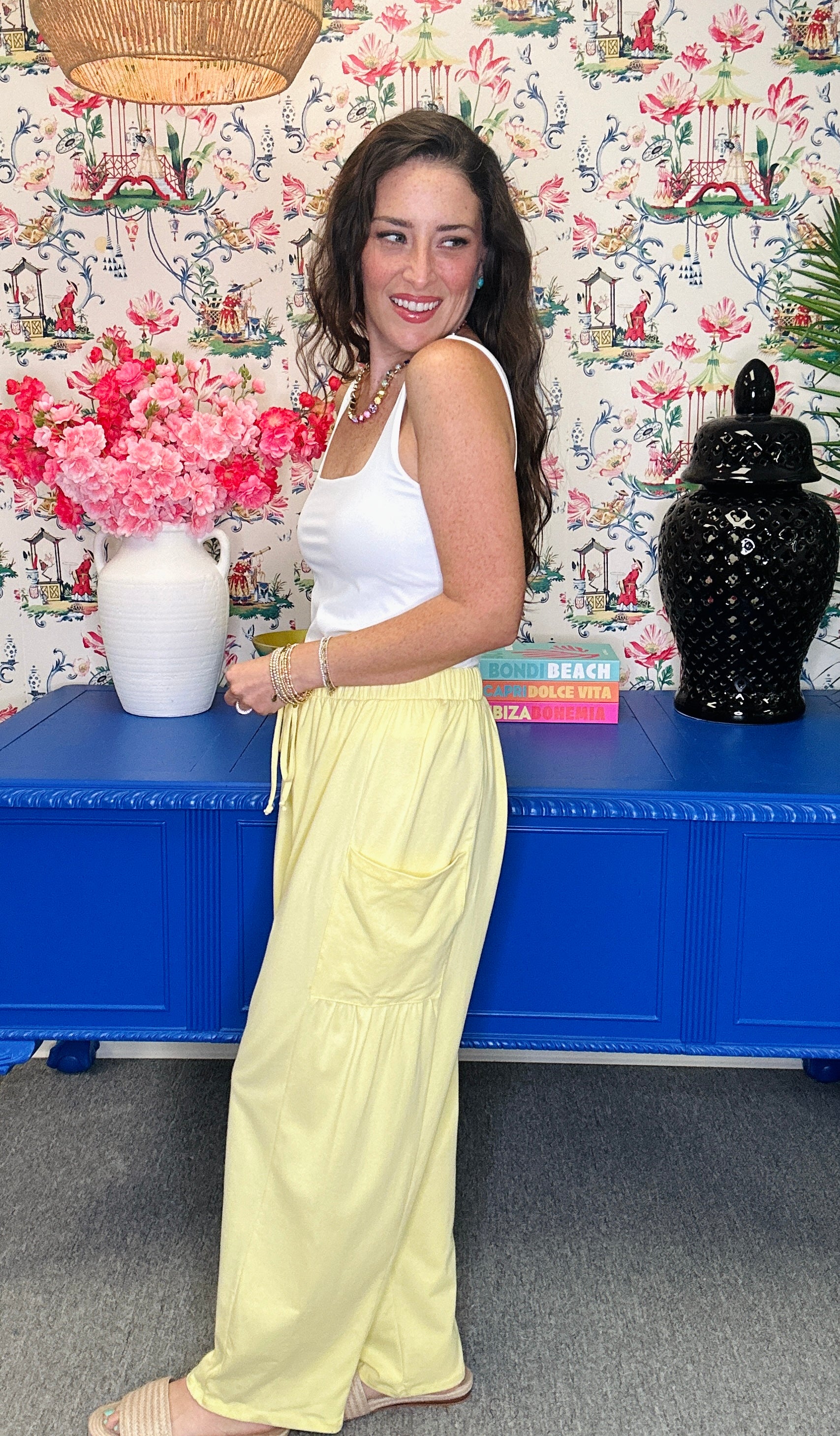 A woman posing in a white tank top and light yellow wide-leg trousers, standing in front of a blue cabinet and a floral wallpaper background.