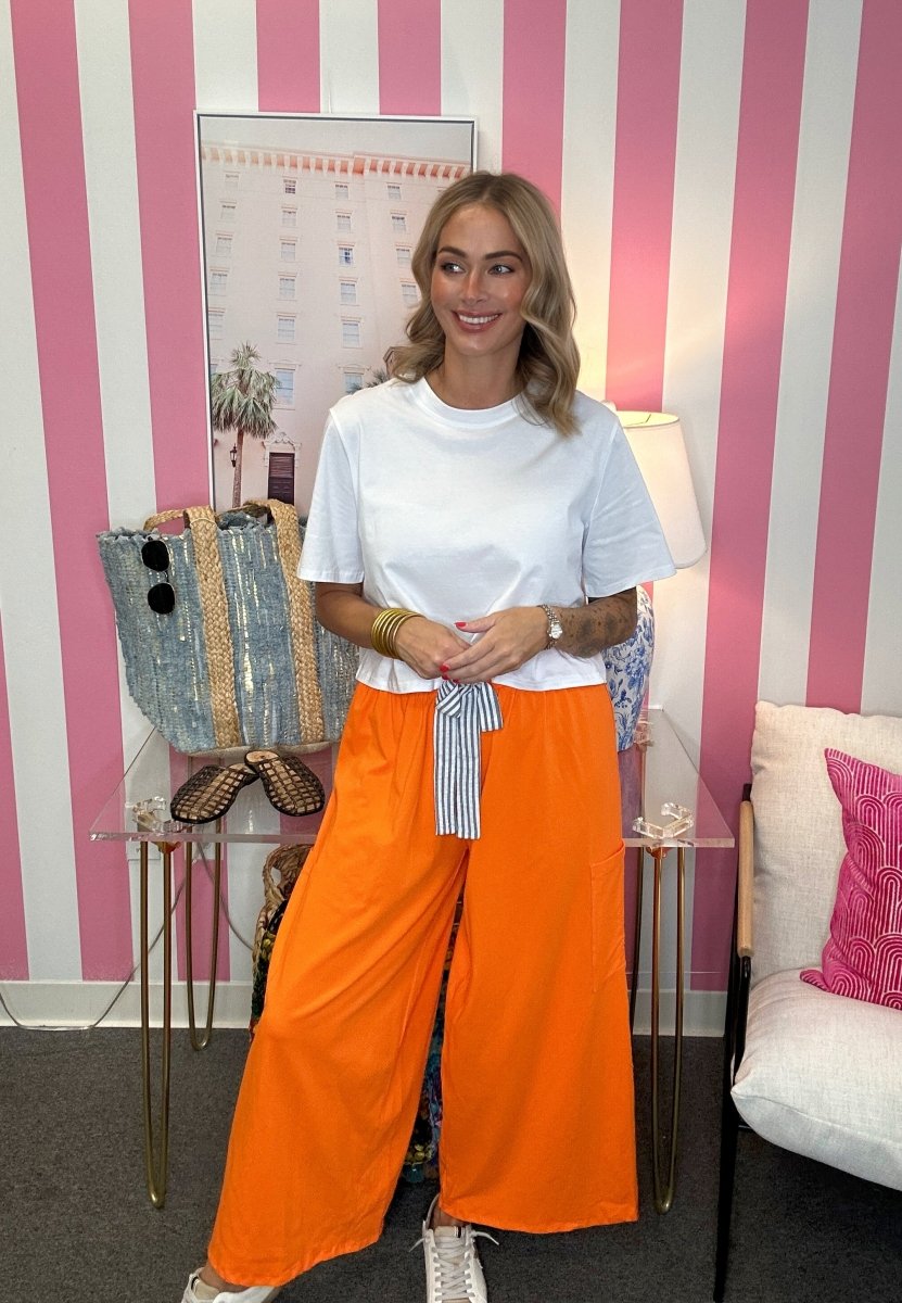 A woman stands in a room with pink and white striped wallpaper, wearing a white crewneck t-shirt and bright orange wide-leg pants with a striped tie belt. She is posing with a denim tote bag on a glass side table.