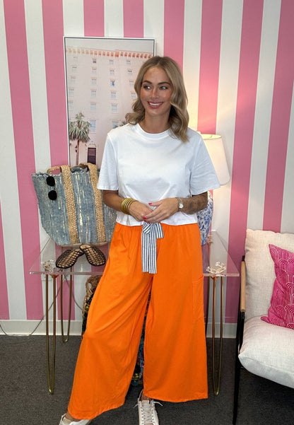 A woman stands in a room with pink and white striped wallpaper, wearing a white crewneck t-shirt and bright orange wide-leg pants with a striped tie belt. She is posing with a denim tote bag on a glass side table.