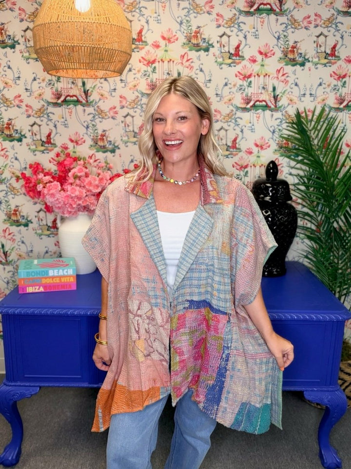 A smiling blonde woman stands indoors wearing a loose, patchwork-style button-up shirt in soft pastel tones with blue plaid and pink floral sections, paired with light blue jeans. She is posed in front of floral wallpaper, a blue side table, pink flowers, and a woven pendant light.