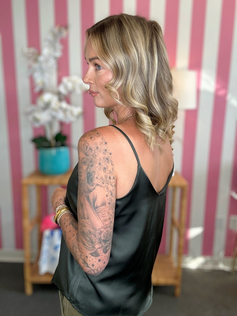 A woman with blonde hair and a large tattoo on her right arm is shown from the back and side, wearing a black satin camisole top in a room with pink and white striped wallpaper.