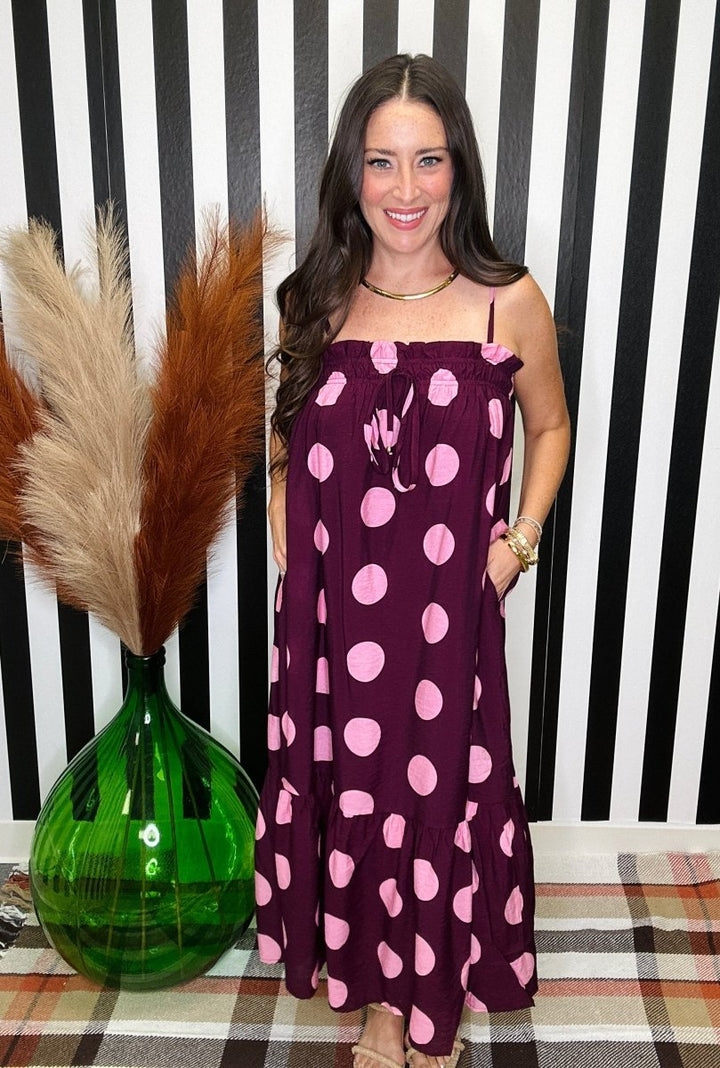 A woman wearing a dark purple spaghetti-strap maxi dress with large pink polka dots, standing in front of a black and white striped wall next to a green glass vase with pampas grass.