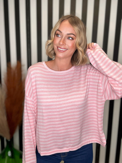 A woman with blonde wavy hair wearing a pink and white striped long-sleeve crop top and blue jeans, posing in front of a black and white striped background.