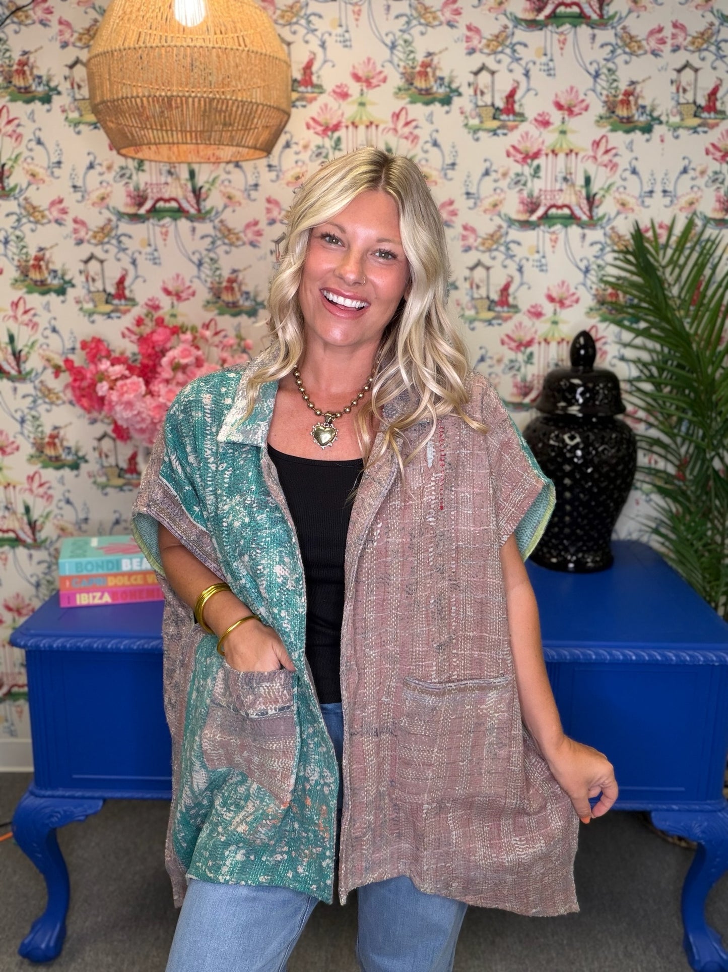 A woman modeling a two-piece linen set consisting of a black top and a matching long vest in a bohemian-themed room with floral wallpaper, a blue table, and a woven lamp.