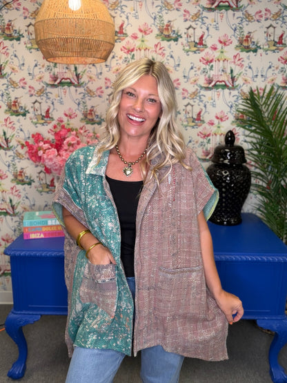 A woman modeling a two-piece linen set consisting of a black top and a matching long vest in a bohemian-themed room with floral wallpaper, a blue table, and a woven lamp.