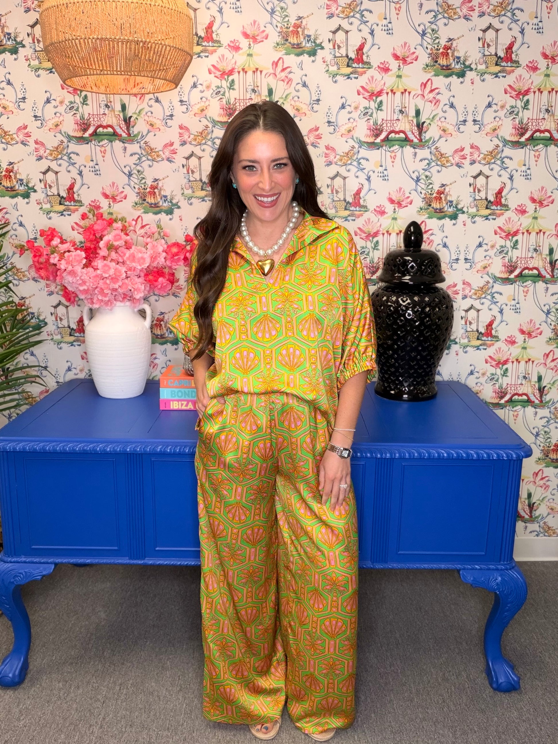 Woman in a colorful outfit standing in front of a floral-patterned wall with a blue table and decorative items.