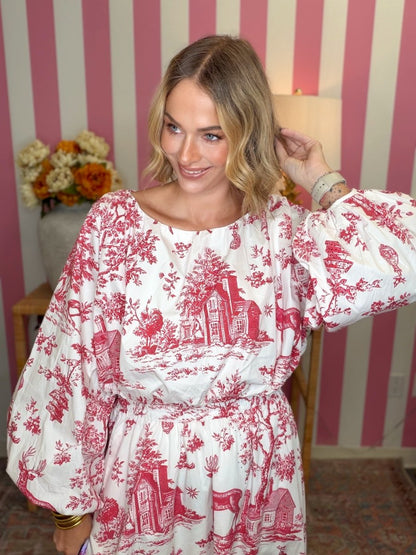 A woman wearing a white dress with a red toile pattern, standing in front of a pink and white striped wall.