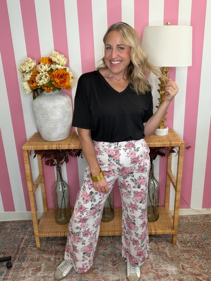 A woman stands in a room with pink and white striped wallpaper, wearing a black short-sleeve V-neck top and white wide-leg pants with a pink floral print. She is posing in front of a wooden side table decorated with a vase of flowers and a lamp.