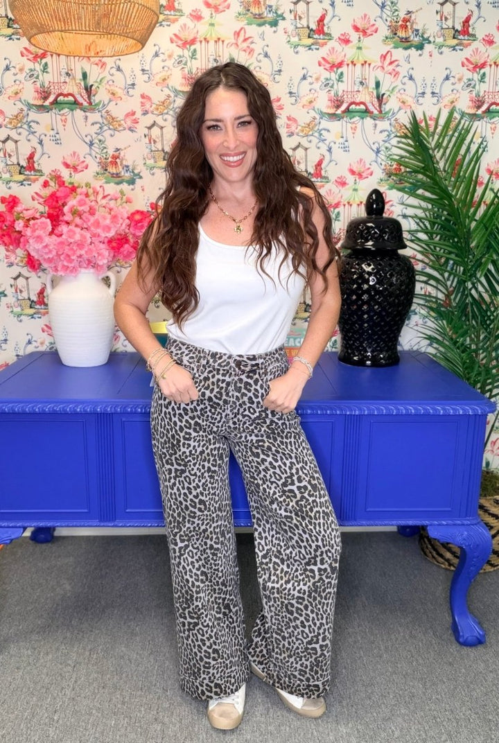 A woman stands indoors wearing a white sleeveless top and black-and-white leopard-print wide-leg pants, posed in front of a blue cabinet with floral wallpaper and decorative objects.