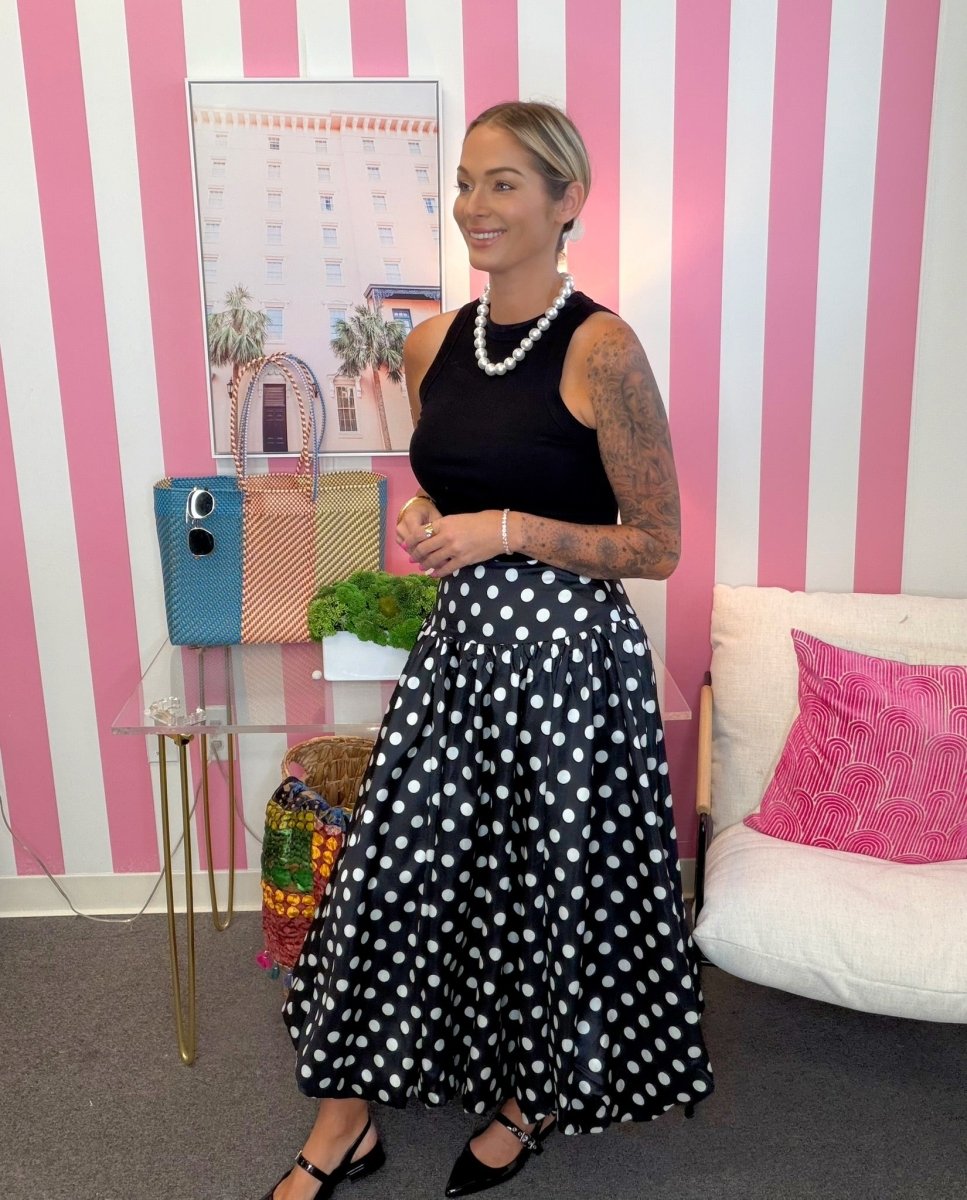 A woman stands in a room with pink and white striped walls, wearing a black sleeveless top and a black midi skirt with white polka dots. She is accessorized with a pearl necklace and black heels.
