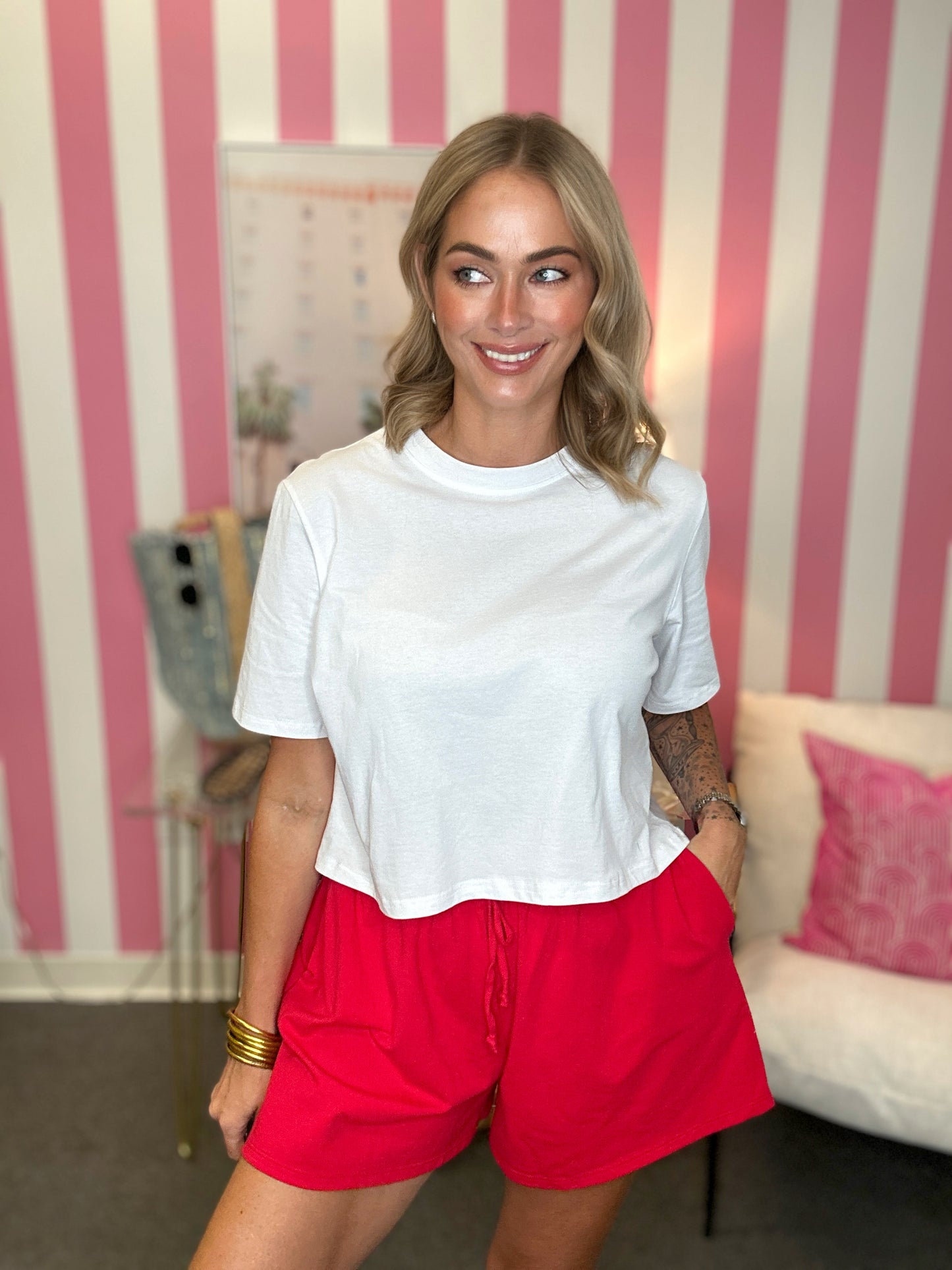 A woman wearing a white short-sleeve crop top and bright red shorts, standing in front of a pink and white striped wall in an indoor room setting.