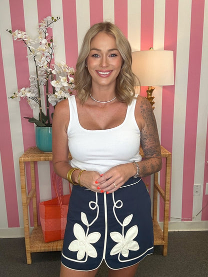 Woman wearing a white tank top and navy skirt with floral patterns in a room with striped walls.